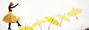 Eine Frau in einem gelben Rock mit einem gelben Regenschirm in der Hand. Sie läuft quasi in der Luft über 6 weitere gelbe Regenschirme.