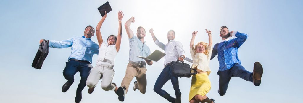 6 Personen, in Businesskleidung, springen in die Luft und freuen sich. Dahinter ein strahlend blauer Himmel